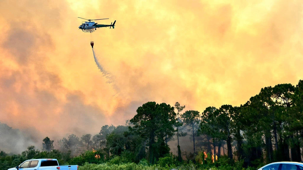 Helicopter dropping water on active Florida wildfire with heavy smoke and flames moving through pine forest during dry conditions